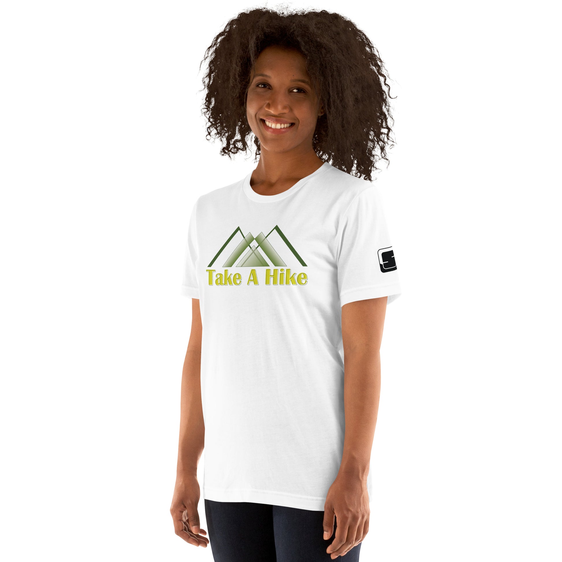 A cheerful African American woman with voluminous curly hair wears a white t-shirt featuring stylized green mountains and yellow text that states “Take A Hike”. She stands confidently, smiling at the camera, her hands slightly behind her hips. The t-shirt's design is clearly visible on her chest, and she has a relaxed posture in front of a white background. A small logo patch is visible on the sleeve of the t-shirt, adding a detail to the simple outdoor-themed attire.