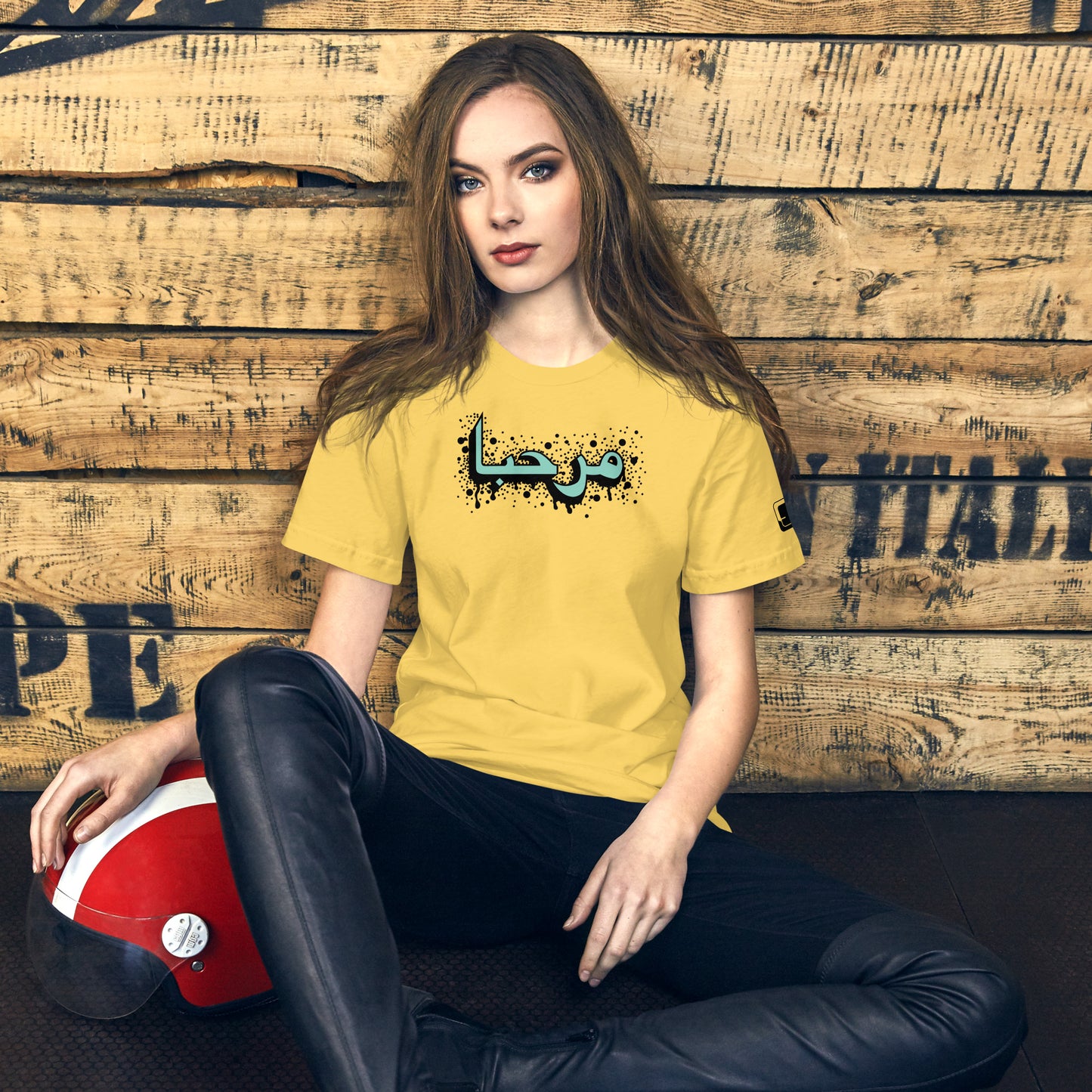 Woman with long hair sitting casually with a red helmet by her side, wearing a mustard yellow t-shirt featuring a turquoise graphic with Arabic calligraphy for "hello", paired with black pants, against a rustic wooden backdrop.