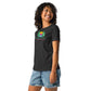 A smiling woman wearing a dark gray t-shirt with a colorful design featuring two green pine trees framing a striped sunset in pink, orange, and yellow. Below the graphic, the text reads "Shady Pines Escape Club" in green and pink. She pairs the shirt with casual denim shorts, creating a relaxed, outdoorsy vibe.