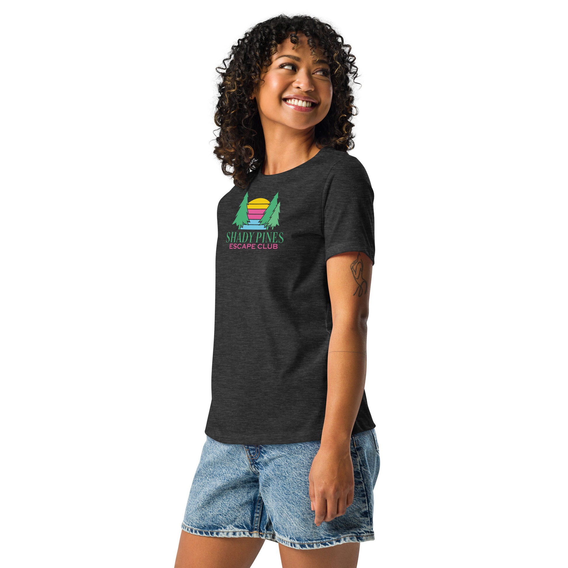 A smiling woman wearing a dark gray t-shirt with a colorful design featuring two green pine trees framing a striped sunset in pink, orange, and yellow. Below the graphic, the text reads "Shady Pines Escape Club" in green and pink. She pairs the shirt with casual denim shorts, creating a relaxed, outdoorsy vibe.